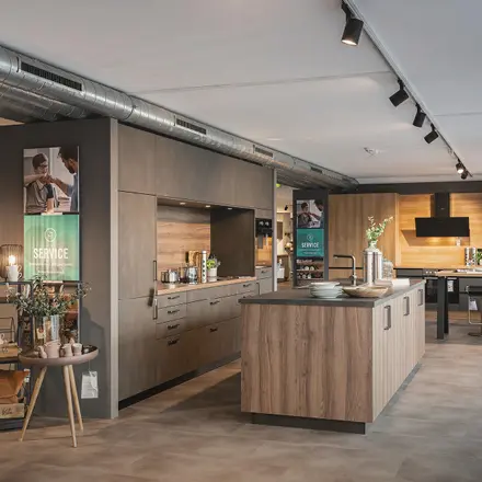 Küchenstudio mit großer Insel in Holzoptik, Spüle, Einbauküche an der Wand und Sitzplatz links im Eingangsbereich.